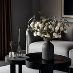 Matte black metal vase with white dried flowers on a round black coffee table in a modern living room.